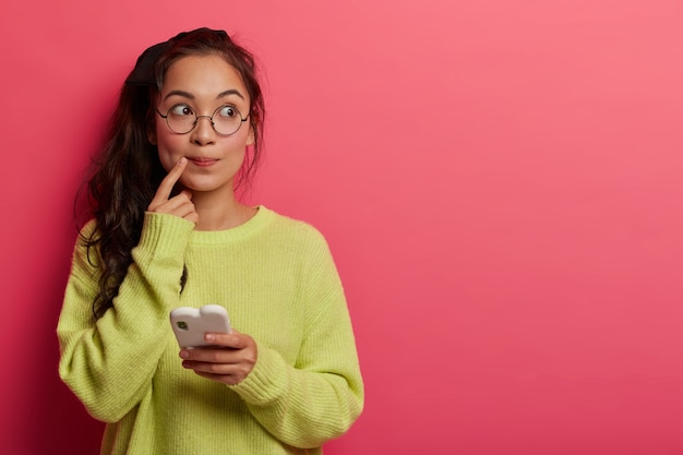 Photo of pensive surprised woman keeps index finger near lips, looks aside, thinks about message content, holds mobile phone