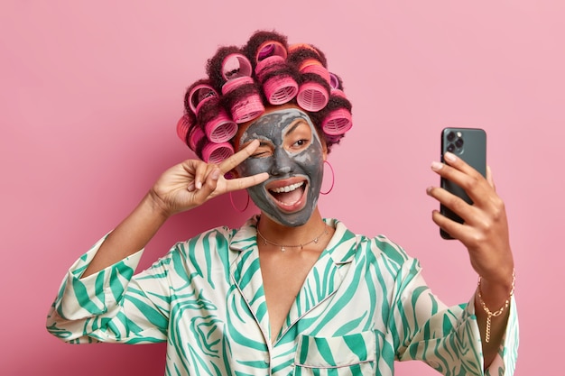 Free photo photo of pleased ethnic woman makes peace gesture over eye smiles broadly applies beauty mask hair rollers takes selfie portrait on mobile phone wears dressing gown isolated over pink wall