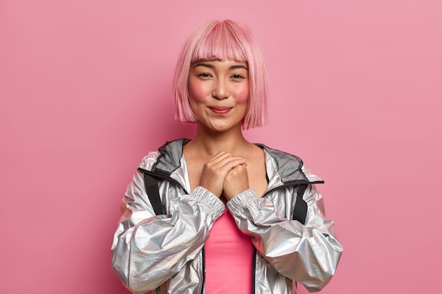 Free Photo photo of pleased smiling shy teenage woman has pink hair wig with fringe, keeps hands pressed together, anticipates for something good happen, dressed in fashionable silver jacket, models indoor