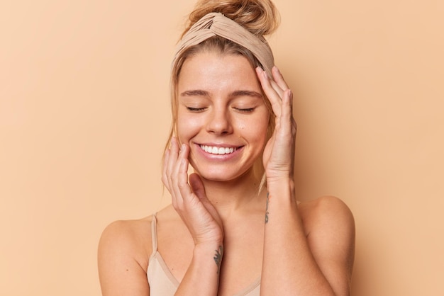 Free Photo photo of pleased young european woman keeps eyes closed touches face gently has natural beauty healthy clean skin wears headband has combed hair isolated over beige background. wellness concept