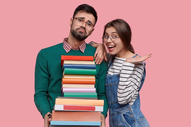 Free photo photo of positive european woman wears striped sweater and overalls, leans at shoulder of tired male nerd with pile of thick books