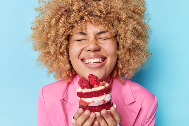 Free photo photo of positive young woman with curly hair holds appetizing cake with raspberries enjoys eating sweet creamy food closes eyes from satisfaction dressed in pink formal jacket eating dessert