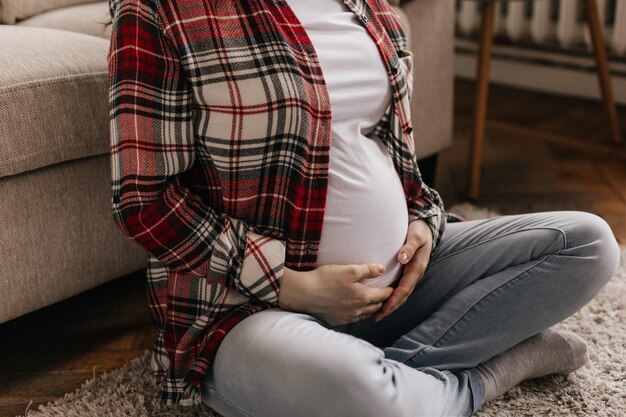 Photo of pregnant woman dressed in denim pants red and black plaid shirt white tee sitting on beige soft carpet near sofa