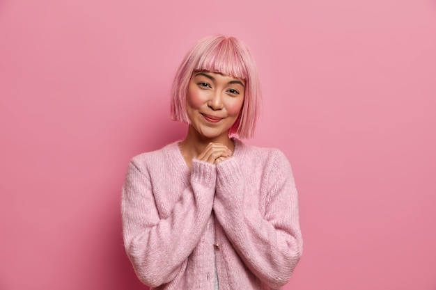 Free photo photo of pretty asian woman in good mood, keeps hands together, has charming smiling expression, thinks about something pleasant wears pink jumper stands indoor.