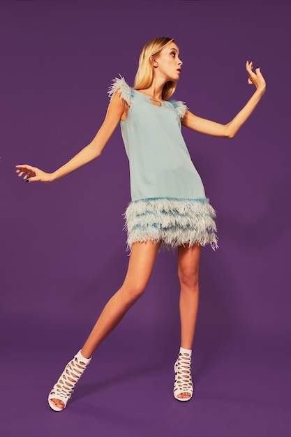 Photo of pretty young blonde girl in blue cocktail dress dancing on purple background in studio. Indoor portrait of caucasian young lady expressing her emotions