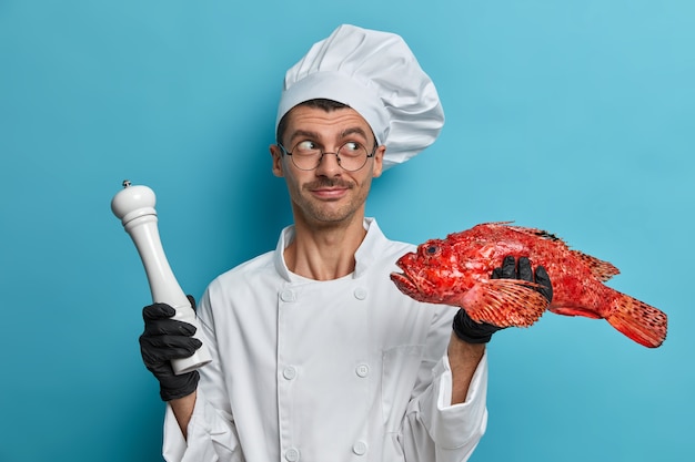 Free photo photo of professional male chef holds raw red sea bass and pepper mill for seasoning, wears cook uniform