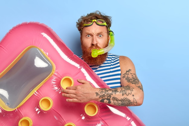 Free photo photo of redhead ginger man wears swim goggles, snorkeling mask, going to dive and swim in sea, holds pink inflated mattress, looks seriously