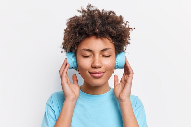 Photo of relaxed curly haired woman closes eyes enjoys music keeps hands on headphones with good sound quality dressed in casual blue t shirt isolated over white wall. Lifestyle concept