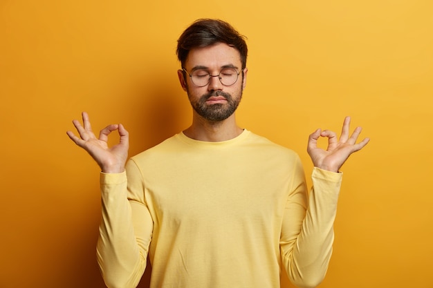 Free photo photo of relaxed unshaven european man stands in lotus pose, makes zen gesture, breathes deeply and tries to relax, keeps eyes closed, wears glasses and jumper, poses indoor, reaches nirvana