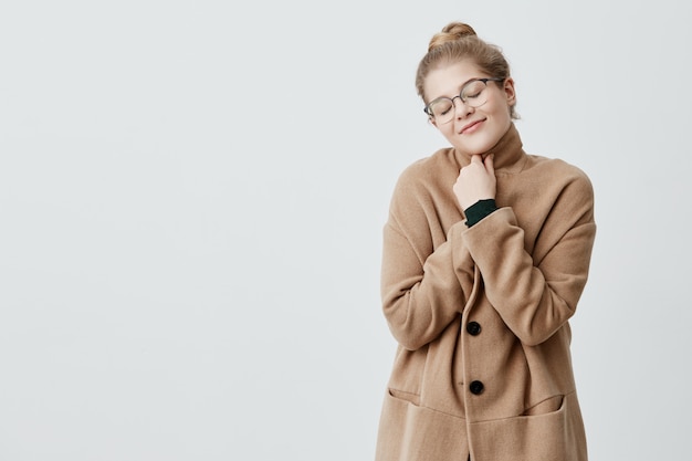 Photo of restful woman with blonde hair in knot wrapping herself in coat having sincere and satisfied smile, closing her eyes in delight posing . Happy female rejoicing life