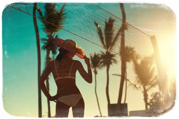 Photo in retro style  of sexy model girl in white bikini with volleyball net on beach and palms behind blue summer sky