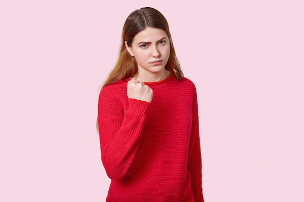 Photo of sad angry young European woman with straight dark hair, shows fist at camera, dressed in red jumper, poses over rosy wall expresses threating, reproaches and promises to revenge.