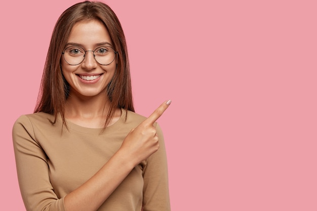 Free photo photo of satisfied brunette caucasian girl with tender smile, satisfied, indicates with index finger at upper right corner