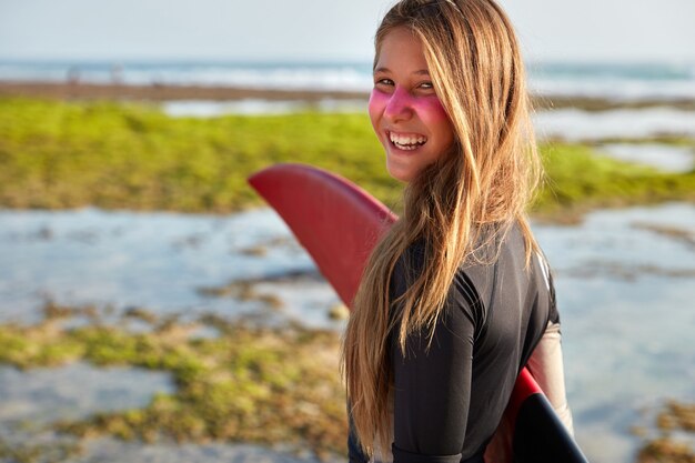 Photo of satisfied long haired woman holds surfboard under hand, looks positively