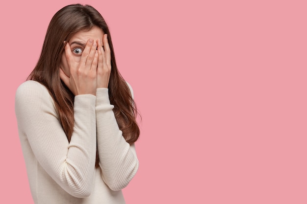 Photo of scared woman covers face with both hands, looks through fingers, peeks with fear, has dark hair, dressed in comfortable jumper