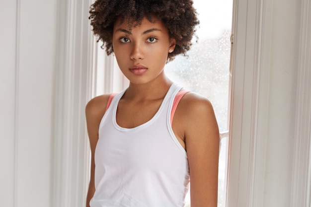 Free photo photo of serious concentrated dark skinned woman with curly hair, dark skin, dressed in casual white t shirt, stands near window