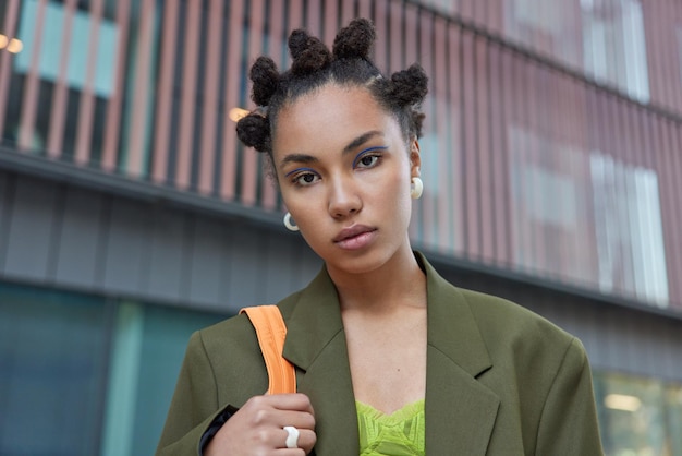 Free photo photo of serious hipster girl with hair buns blue eyeliner dressed in formal dark green jacket looks directly at camera walks outdoors poses listens something attentively