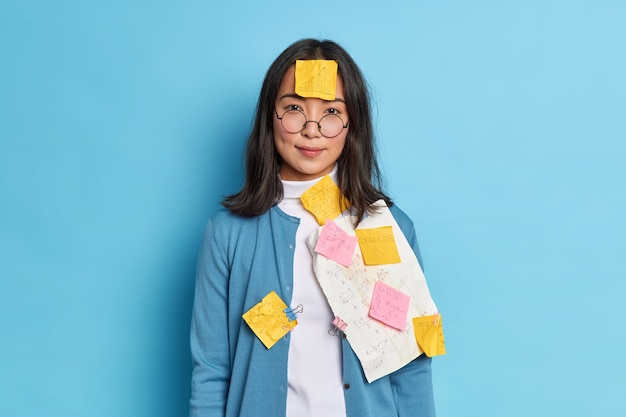 Free photo photo of serious student makes notes on stickers and papers to remember information wears round spectacles prepares for private lesson works on coursework in university.