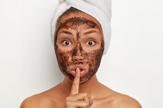 Photo of shocked Afro American woman with coffee scrub for peeling from pores, makes silence gesture, has surprised face expression, shows bare shoulders, tells secret, has cosmetology procedure