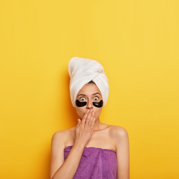 Free photo photo of shocked european woman covers mouth with palm, applies patches under eyes to remove wrinkles, takes bath regualarly, wants to have perfect skin