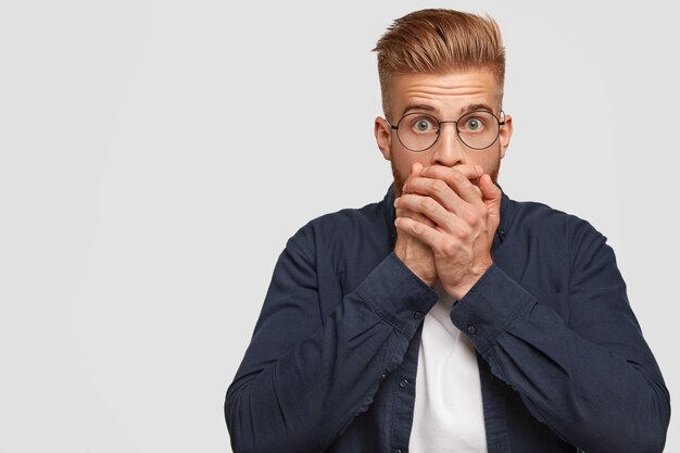 Photo of shocked ginger man has trendy hairstyle, covers mouth with both palms, keeps intriguing information in secret