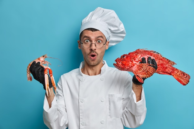 Free photo photo of shocked skilled chef holds crayfish, red bass, cooks delicious dish from seafood, works on his fish menu, keeps mouth opened with wonder