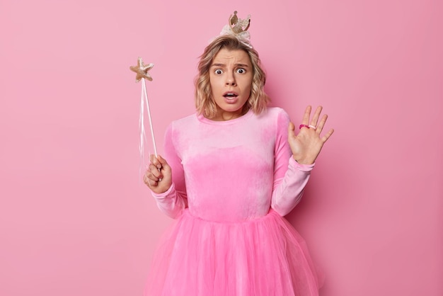 Photo of shocked young pretty woman fairy wears pink dress and crown holds magic wand stares with frightened expression cannot believe her eyes poses indoor prepares for party and celebration