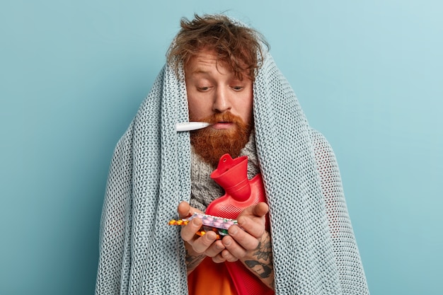 Photo of sick ginger man feels sick, has winter flu virus, suffers from fever and high temperature, holds thermometer in mouth, wrapped in coverlet, warms with hot bottle, has different medicaments