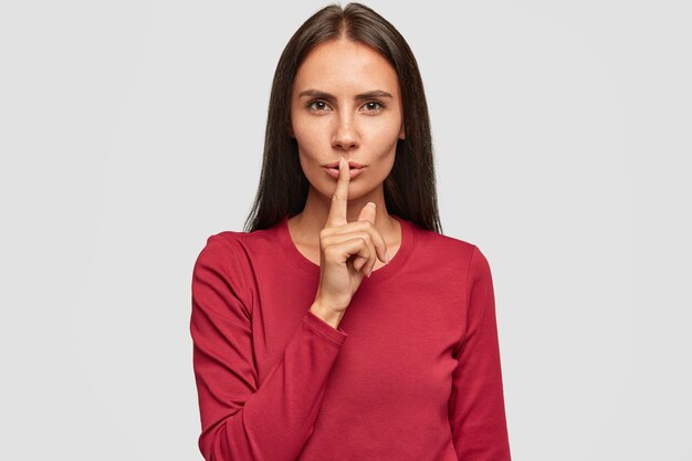 Photo of silent beautiful brunette lady dressed in red sweater, keeps fore finger on lips, has serious expression,