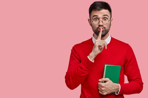 Photo of silent male teacher keeps index finger over mouth, asks pupils not make noise during lecture, holds notebook with notes