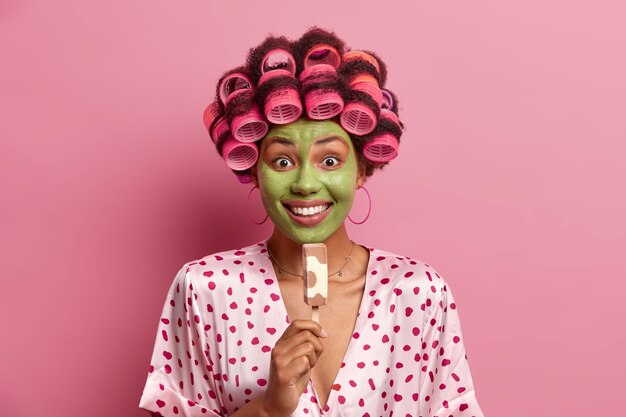 Photo of smiling woman smiles broadly, applies green moisturizing mask, eats delicious ice cream on stick, wears hair curlers, silk dressing gown, enjoys summer time. Housewife with frozen dessert