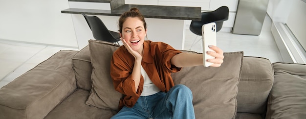 Free Photo photo of smiling young woman sitting on couch posing on sofa in living room taking selfies for