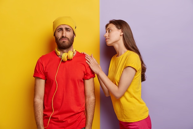 Free photo photo of sorrowful unshaven man has trouble, wears yellow hat and red t shirt, smirks face with displeasure, caring girlfriend touches his shoulder, tries to calm and help in difficult situation