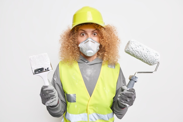 Free photo photo of stunned curly female engineer holds painting roller and brush going to redecorate house dressed in building uniform paints interior wall of new apartment