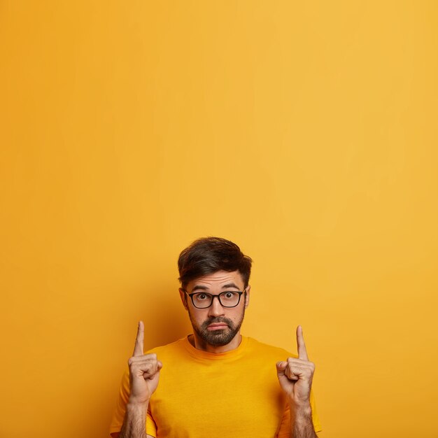 Free Photo photo of stunned man shows product, recommends service with amazed expression, stares with bugged eyes, indicates on blank space upwards, wears optical glasses, isolated on yellow wall