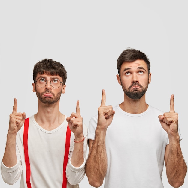 Free Photo photo of sullen men with dark hair, dressed in white clothes, points upwards, have displeased facial expressions