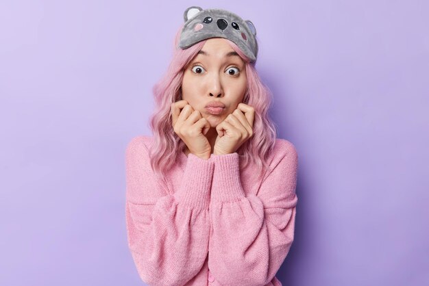 Photo of surprised Asian woman with healthy skin keeps hands under chin has folded lips widely opened eyes reacts on something shocking wears sleepmask and pink jumper isolated over purple background