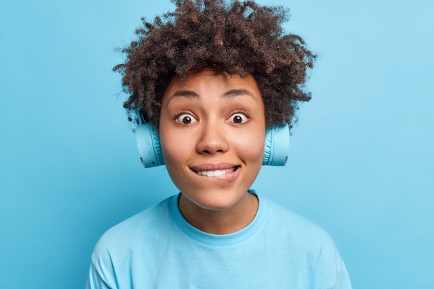 Free photo photo of surprised curious afro american woman bites lips looks directly  wears wireless headphones dressed in casual t shirt isolated over blue wall. people leisure lifestyle