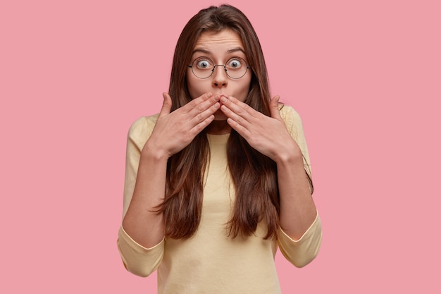 Free photo photo of surprised dark haired woman covers mouth, looks with frightened expression, reacts at something astonishing