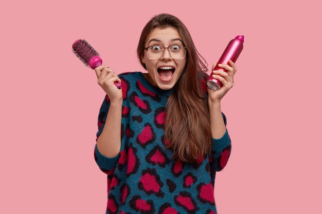 Free Photo photo of surprised european woman keeps mouth widely opened with positive expression, holds spray and hairbrush, has overjoyed look at mirror