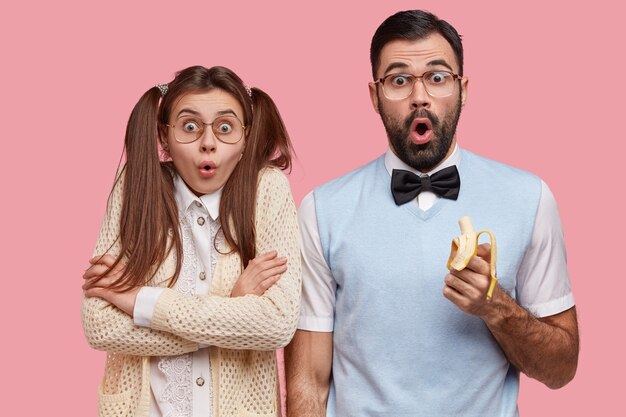 Photo of surprised female and male wonks stare with disbelief, eat delicious banana, dressed in old fashionable clothes