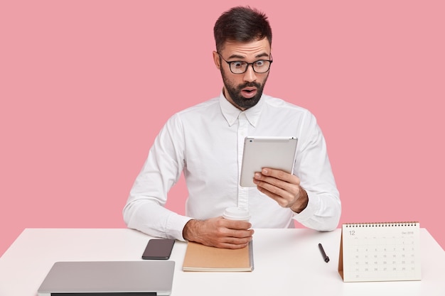 Free photo photo of surprised male employee stares at screen of touchpad, feels astonishment, reads shocking message, drinks coffee from disposable cup, wears spectacles