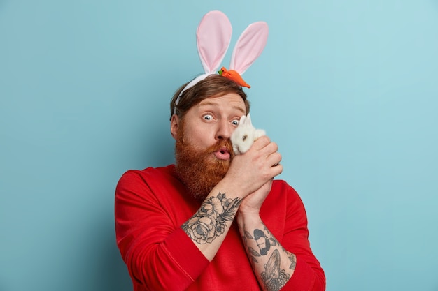 Free photo photo of surprised redhead man keeps small animal near face, plays with white rabbit, wears bunny ears, prepares for costume party on easter eve, poses against blue wall. spring holiday