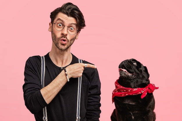 Photo of surprised young male pet lover points at his pedigree dog, dressed in black shirt, wonders something, indicates index finger for paying attention to animal, isolated over pink wall.