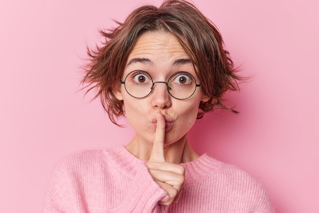 Free photo photo of surprised young woman with short hairstyle makes hush gesture asks to keeps quiet presses index finger over lips wears round transparent glasses and casual jumper poses indoor. do not speak