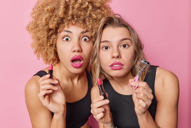 Free photo photo of surprised young women apply lipstick hold cosmetic products do makeup stare shocked at camera want to look beautiful dressed casually isolated over pink background beauty procedures