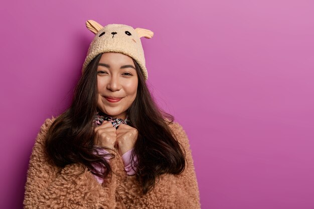 Photo of teenage girl wears fashionable hat and brown coat, looks sincerely with gentle smile, enjoys wearing new outfit