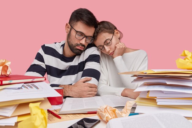 Photo of tired overworked sleepy young woman and man keep heads near, look fatigue, wear optical spectacles, read information from encyclopedia