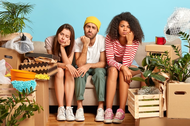 Free photo photo of tired three diverse students move in new flat, change place of living, have sleepy dissatisfied expressions, fatigue after unpacking belongings, surrounded with items in carton containers