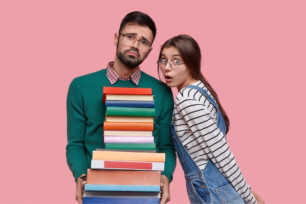 Free photo photo of tired unshaven man with pity expression, holds pile of books, looks in displeasure, his female classmate has stupefied look